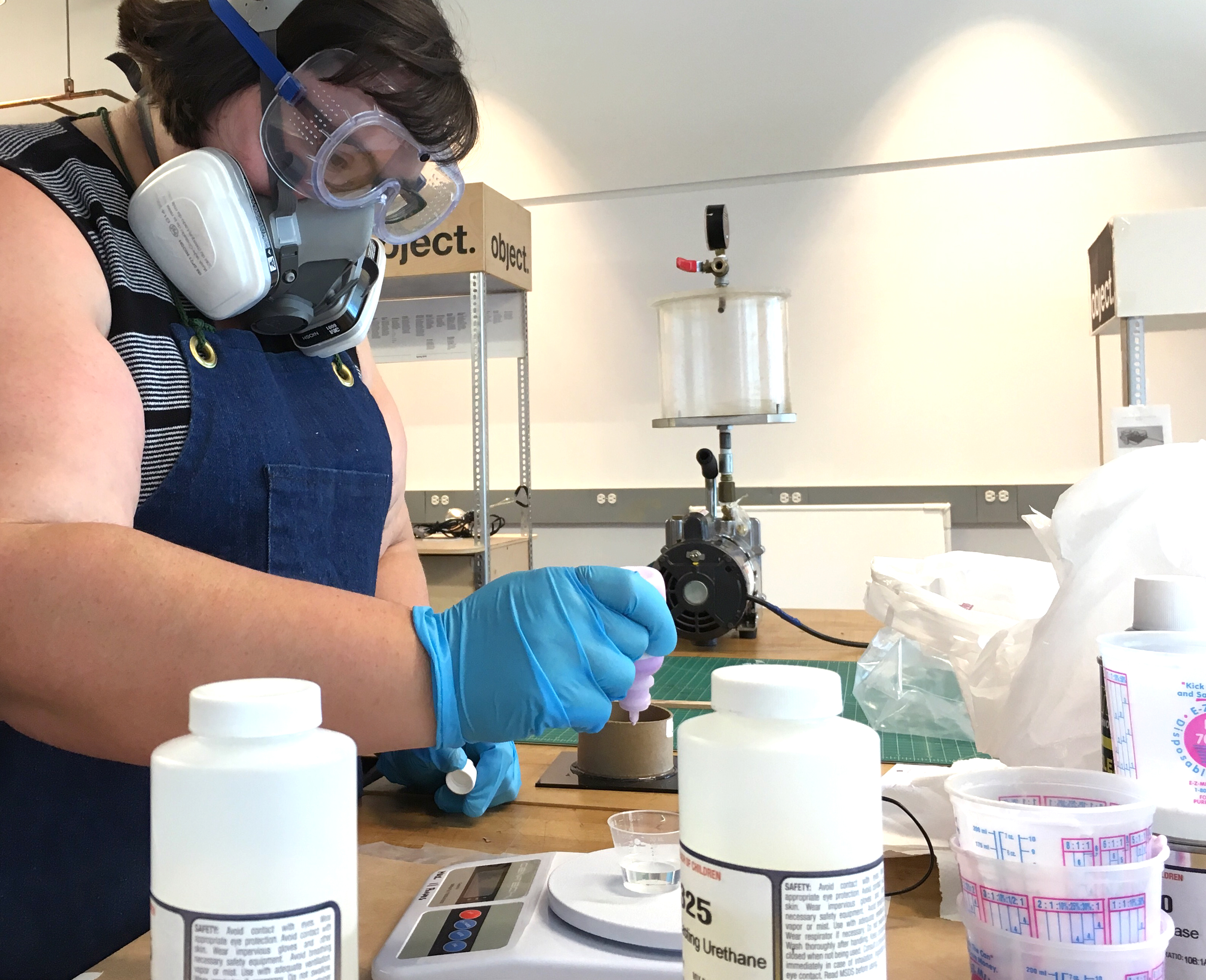 A fat white woman with brown hair wears safety goggles, a half-mask respirator, and blue nitrile gloves. She's pouring drops of liquid into a small container on a digital scale. She's surrounded by mold-making accessories, like bottles of urethane, cardboard molds, plastic cups, and a vacuum degasser.